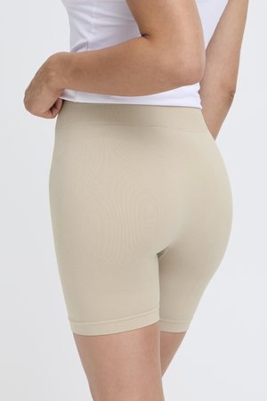 Femme portant un short beige moulant sans coutures et un haut blanc sans manches, vue de dos sur un fond uni.