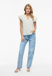 Top gris claro con mangas cortas acampanadas, escote en V, combinado con pantalones vaqueros holgados azules y sandalias de tacón beige. Tejido suave y ajuste casual.