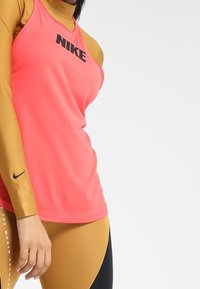 Nike pink ärmelloses Top mit schwarzem Logo, kombiniert mit einem goldenen Langarm-Basisschicht und schwarzen Leggings mit einem geschwungenen Farbblockdesign.