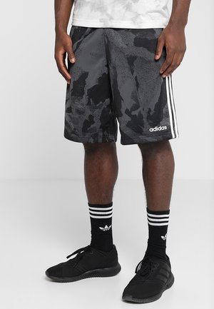 Mand iført sorte og grå camouflage Adidas shorts, sorte Adidas sokker med hvide striber og sorte sportssko.