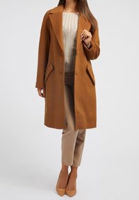 Bruine wollen jas met een notch-kraag, twee voorzakken en knopen. Gecombineerd met een beige top en broek, gedragen met beige hoge hakken.