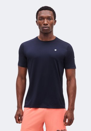 Mand iført en tætsiddende mørkeblå t-shirt og ferskenfarvede shorts, stående mod en ensfarvet lys baggrund, vendt fremad.