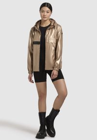 Metallisch goldene Kapuzenjacke mit Reißverschluss vorne, elastischen Bündchen und einer Brusttasche mit einem schwarzen Markenlabel; kombiniert mit dunklen Shorts und schwarzen Stiefeln.