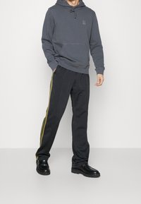 Sudadera gris con bolsillo frontal y capucha ajustable, combinada con pantalones negros que tienen rayas laterales amarillas y verdes, y calzado negro.