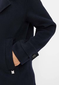 Manteau en laine marine avec un col cranté, des poignets boutonnés, des poches latérales et un accent logo en métal sur l'ourlet. Texture douce et design élégant.