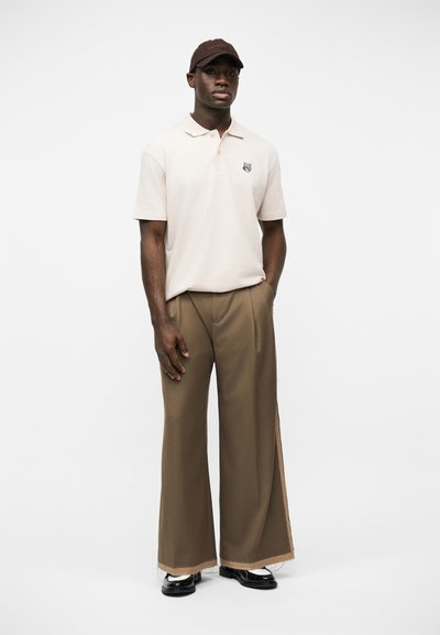 Άνδρας που φοράει μπεζ πόλο πουκάμισο με μικρό έμβλημα, φαρδιά καφέ παντελόνια, μαύρα παπούτσια και σκουρό καφέ καπέλο μπέιζμπολ, στέκεται μπροστά σε λευκό φόντο.