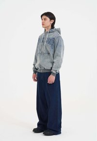 Šedá džínová mikina na zip s modrým výšivkovým textem, v kombinaci s volnými tmavě modrými širokými kalhotami a černými nazouvacími botami.