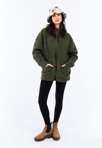 Veste en polaire surdimensionnée vert olive avec poches avant, associée à un legging noir et des bottines beige. Un chapeau seau à motifs blancs ajoute une touche de détail.