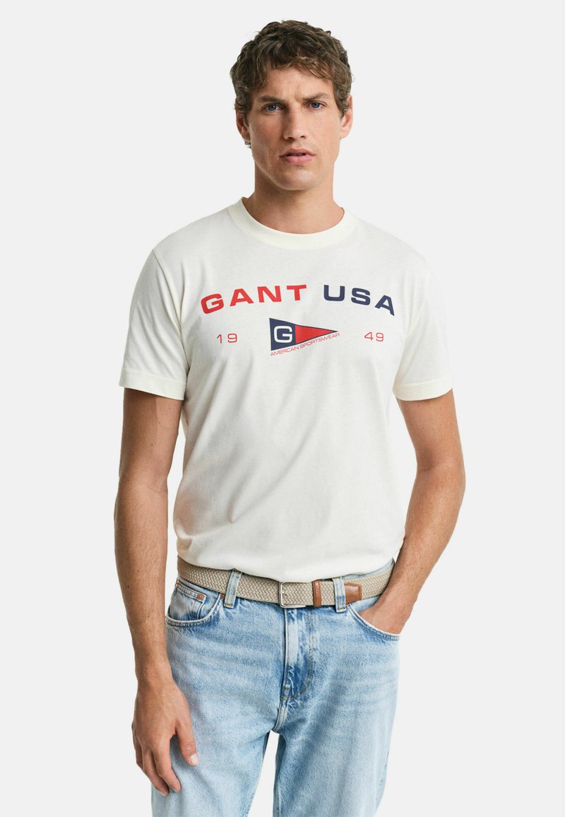 Krémové tričko s krátkým rukávem s červeným a tmavě modrým textem "GANT USA" a trojúhelníkovým logem, kombinované se světlomodrými džíny a tkaným opaskem.