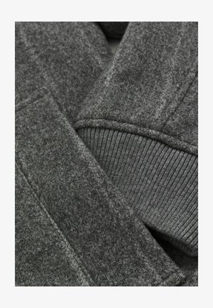 Prim-plan al unui manșon de palton gri din lână, cu un tiv tricotat în rânduri și detalii vizibile ale cusăturii și buzunarului.