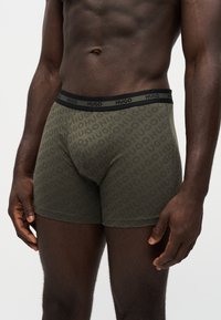 Torse et parties supérieures des jambes masculines portant un boxer vert olive avec le logo "HUGO" répété et une ceinture noire avec le texte "HUGO".
