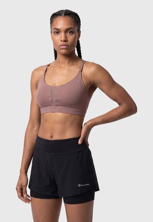 Athletische Frau mit geflochtenem Haar trägt einen mauvefarbenen Sport-BH und schwarze geschichtete Champion-Shorts, steht mit einer Hand auf der Hüfte vor grauem Hintergrund.