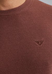 Bordeauxfarbener, strukturierten Pullover mit rundem Ausschnitt und gerippten Bündchen. Besitzt ein dezentes gesticktes Logo, das ein Wabenstrickmuster zeigt.