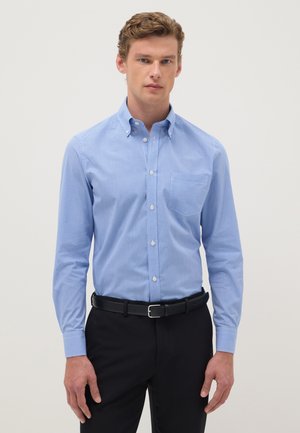 Jeune homme portant une chemise à manches longues à carreaux bleus clairs, rentrée dans un pantalon noir avec une ceinture noire, debout devant un fond uni.