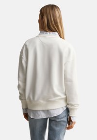 Femme aux longs cheveux blonds portant un sweat-shirt blanc par-dessus une chemise rayée et un jean bleu, de dos sur un fond uni.
