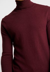 Burgundy polokrage tröja i ribbad stickad textur med långa ärmar, figurnära design och hög hals för extra värme.