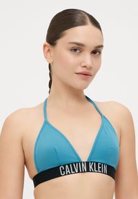 Triangelbikiniöverdel, tillverkad av slätt blått tyg, med ett svart band med "CALVIN KLEIN" i vit text. Sömnadsdetaljer på kuporna.