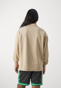 Beige Langarmhemd mit strukturierten Oberflächen, das einen Kragen und einen kontrastierenden Besatz aufweist. Kombiniert mit schwarz gestreiften Shorts und grünen Akzenten.