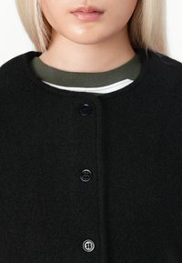 Černý kabát z vlněné směsi s kulatým výstřihem, který je zdobený čtyřmi černými knoflíky. Pod ním se zjevuje zeleno-bílý pruhovaný svetr.
