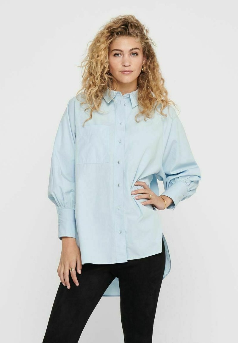 Camicia di jeans azzurra con maniche lunghe e polsini arricciati. Presenta una tasca sul petto e un orlo posteriore leggermente più lungo.