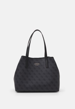 Schwarze Guess Tragetasche mit Monogramm-Muster und zwei langen Henkeln auf schlichtem hellgrauen Hintergrund.
