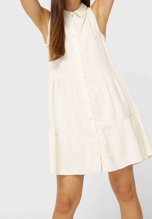 Robe beige sans manches boutonnée avec col et jupe à volants portée par une personne aux longs cheveux bruns, les bras levés au-dessus de la tête.