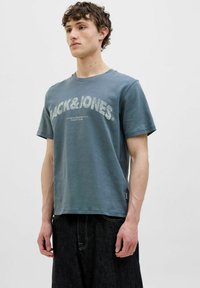 Ubleget blå bomulds t-shirt med "PACK&JONES" logo i lys grå tryk. Rund hals, korte ærmer og et lille mærke ved kanten.