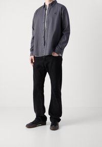 Harmaa nappipaita vaaleanharmaan T-paidan päällä, yhdistetty mustiin housuihin ja tummiin tennareihin, esitellen rentoa istuvuutta ja rentoa tyyliä.