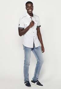 Camisa blanca de botones con mangas cortas y puños de denim, combinada con jeans azul claro y zapatillas de color marino. Presenta botones de metal y detalles de bordado.