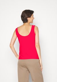 Top vermelho sem mangas com decote arredondado nas costas, feito de tecido suave. Usado com calças bege, apresentando um detalhe impresso subtil na manga.