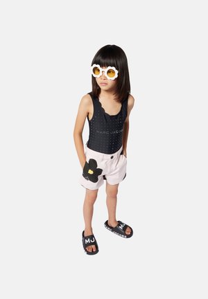 Costume da bagno intero nero con bordi scallop, pantaloni corti rosa con un motivo floreale nero e sandali neri con testo. Occhiali da sole a forma di nuvola bianchi.