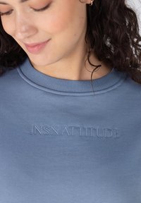 Lichtblauw sweatshirt met ribgebreide kraag en reliëftekst "JNSN ATTITUDE" over de borst. Zachte stofstructuur en casual ontwerp.