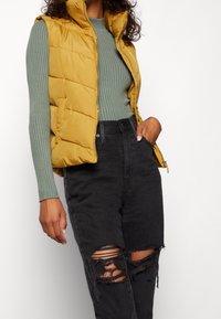 Žlutá péřová vesta s vysokým límcem, na zip, vrstvená přes zelené dlouhé tričko s žebrovaným vzorem a černé distressed džíny.