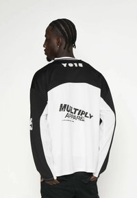 Schwarz-weiße Langarmshirt mit dem Aufdruck 'MULTIPLY APPAREL' und 'YO15' auf dem Rücken. Es hat kontrastierende Ärmel und einen gerippten Kragen.