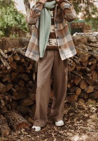 Kurtka w kratę w kolorach beżowym, brązowym i szarym, zielony dzianinowy szalik, brązowy sweter z dzianiny, brązowe plisowane spodnie i białe tenisówki.