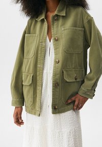 Olivenfarvet oversized jakke lavet af bomuld, med knaplukning, to brystlommer og lange ærmer. Har synlige syningsdetaljer.
