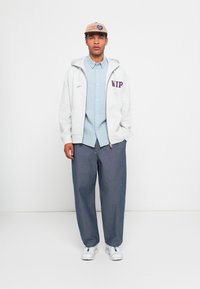 Gray zip-up hoodie with "WIP" in purple, light blue button-up shirt, wide dark blue denim pants, and white sneakers. Beige cap with logo.