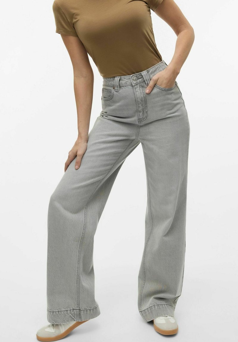 Jeans grises de pierna ancha en tela de mezclilla con cintura alta, que cuentan con bolsillos frontales y costuras visibles. Combinados con zapatillas deportivas beige.