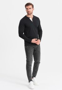 Mies, der ein schwarzes langärmliges Henley-Shirt, eine ausgewaschene graue Jeans und weiße Sneaker trägt, steht mit gefalteten Händen und schaut nach rechts.