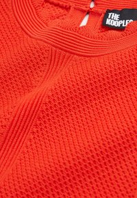Tissu tricoté texturé orange vif avec des détails côtelés et une étiquette noire portant l'inscription « THE KOOPLES ».