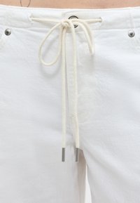 Κοντινό πλάνο λευκού παντελονιού με κορδόνι στο μπροστινό μέρος δεμένο σε φιόγκο, με μεταλλικά τελειώματα και ασημένιες λεπτομέρειες κοντά στη μέση.