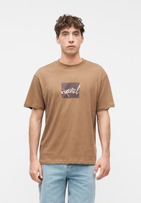 Νέος άνδρας με σγουρά μαλλιά που φοράει καφέ μπλούζα με τετράγωνο γραφικό και λευκό κειμενογράφημα, συνδυασμένη με ανοιχτό μπλε τζιν, που στέκεται μπροστά σε λευκό φόντο.