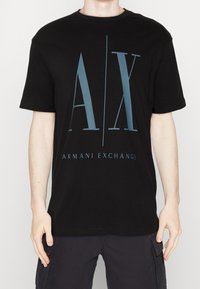 Person som bär en svart Armani Exchange t-shirt med stora blå "A|X" bokstäver och texten "ARMANI EXCHANGE" nedanför på framsidan.
