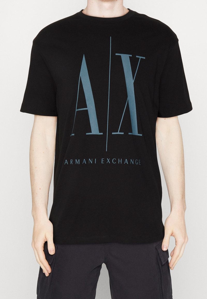 Person som bär en svart Armani Exchange t-shirt med stora blå "A|X" bokstäver och texten "ARMANI EXCHANGE" nedanför på framsidan.