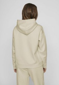 Sudadera beige con una capucha grande, hombros caídos y puños acanalados. Material textil suave, corte relajado y diseño minimalista.