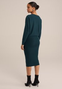 Zelena rebrasta pletena obleka z dolgimi rokavi, v-izrezom na hrbtu in oprijetim krojem; v kombinaciji z črnimi gležnjarji. Gladka tekstura po celotni obleki.