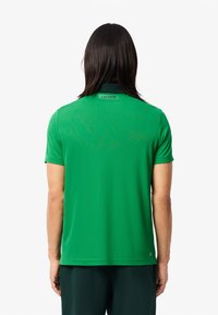 Grünes Poloshirt mit kurzen Ärmeln und einem schwarzen Kragen, mit einem Lacoste-Logo auf der Rückseite. Hergestellt aus glattem, leichtem Material.
