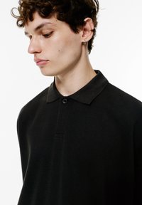 Schwarzes Poloshirt mit klassischem Kragen, kurzen Ärmeln und Knopfleiste. Der Stoff wirkt glatt mit minimaler Textur.
