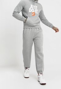 Conjunto de sudadera y pantalones deportivos grises, hecho de una tela suave, con un gráfico blanco de "JUST DO IT" y el logo de Nike en naranja, terminado con puños elásticos.