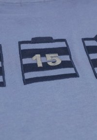 Tissu bleu avec un gros plan de l'icône de calendrier brodée montrant le numéro 15 en beige sur des rayures bleu foncé et bleu clair.